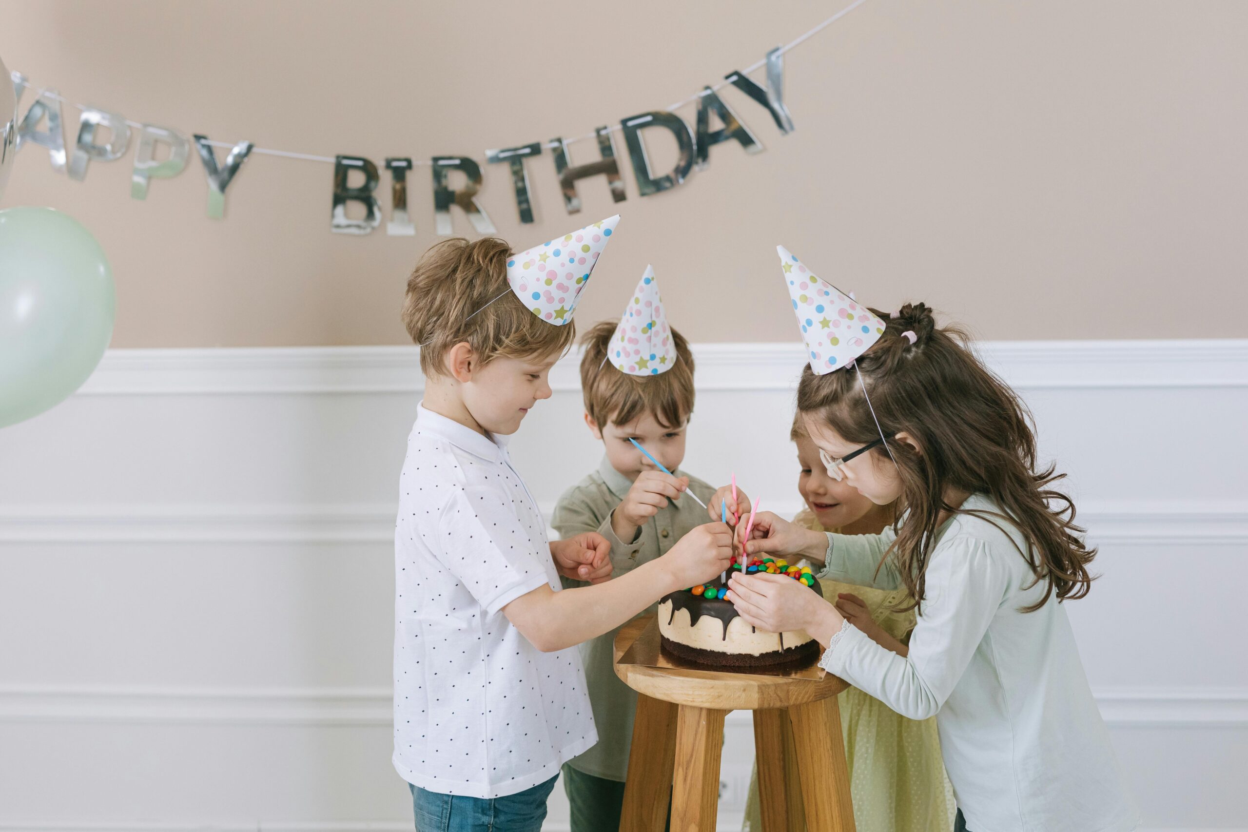 Children in party hats gather around a birthday cake indoors with a 'Happy Birthday' banner.